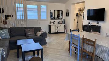 Een woonkamer met een bank, salontafel en poef, een eethoek met een tafel en stoelen, een aan de muur bevestigde televisie, en een keuken met kastjes en een aanrecht.