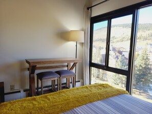 A bedroom with a bed, a desk with two chairs, a lamp, and a large window with a view of a forest and buildings. - Copper Mtn. Studio Queen+Pull out ski in ski out (Copper Mountain)