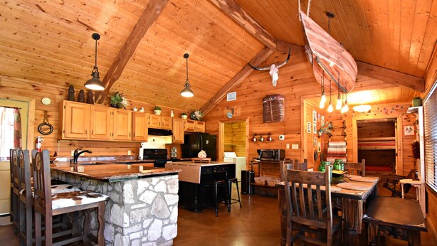 A rustic cabin-style kitchen with wooden cabinets, a stone island, and a dining area with wooden chairs. - Rustic lodge retreat minutes from the Frio! (Concan)