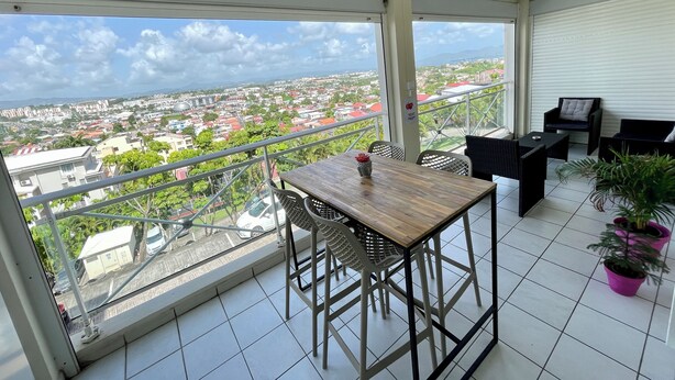 Un balcón con mesa y sillas de madera, plantas en macetas y vista a un paisaje urbano.
