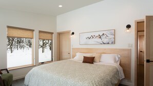 A bedroom with a large bed, two windows with blinds, a painting of birds, and a door to another room.