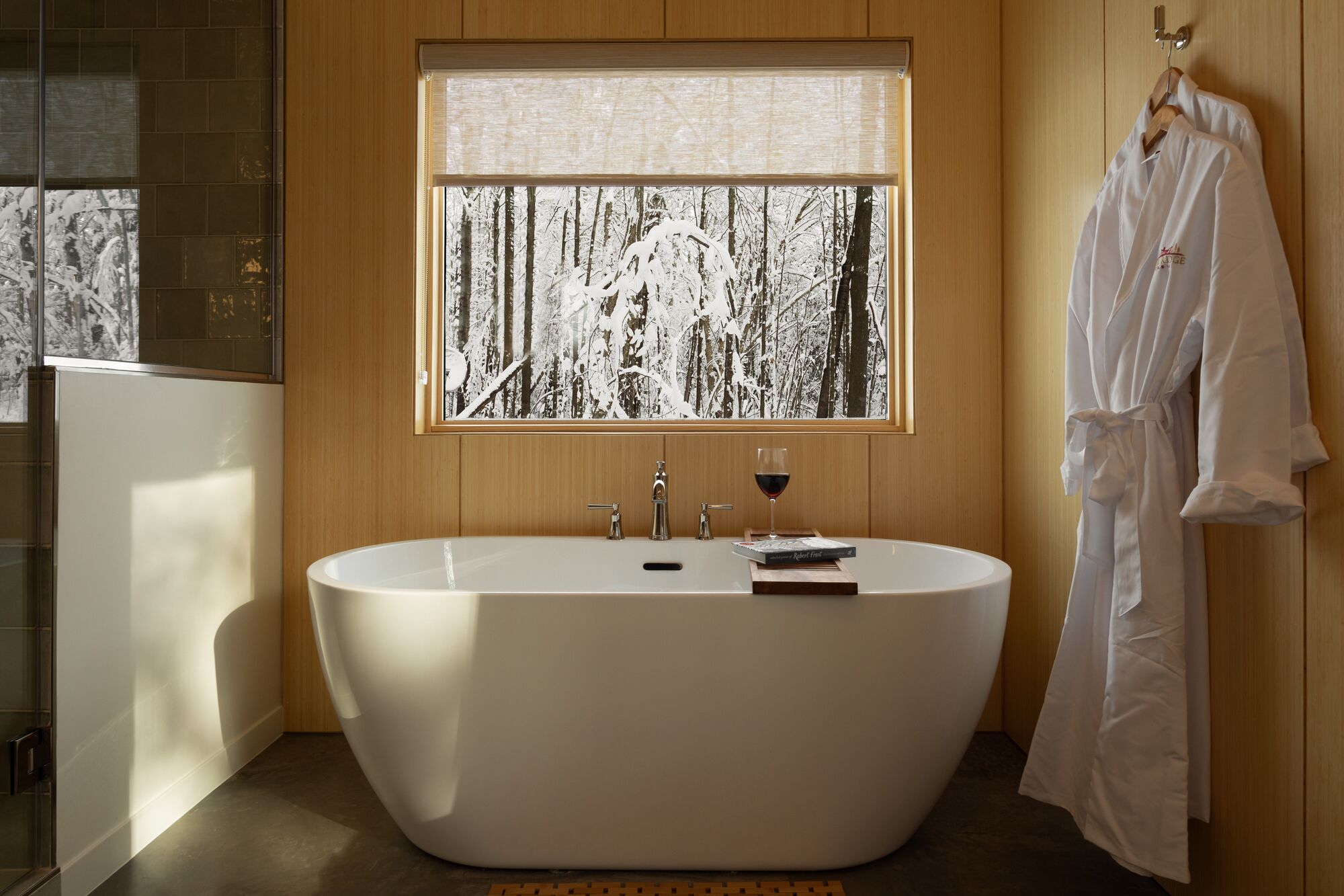 Un baño moderno con una bañera independiente, un ventanal con vistas a un bosque nevado y una bata colgada en la pared.