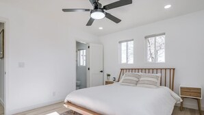 A bedroom with a bed, a ceiling fan, two windows, a wooden nightstand, and a door leading to a bathroom.