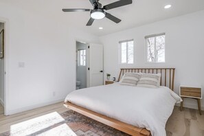 A bedroom with a bed, a ceiling fan, two windows, a wooden nightstand, and a door leading to a bathroom.
