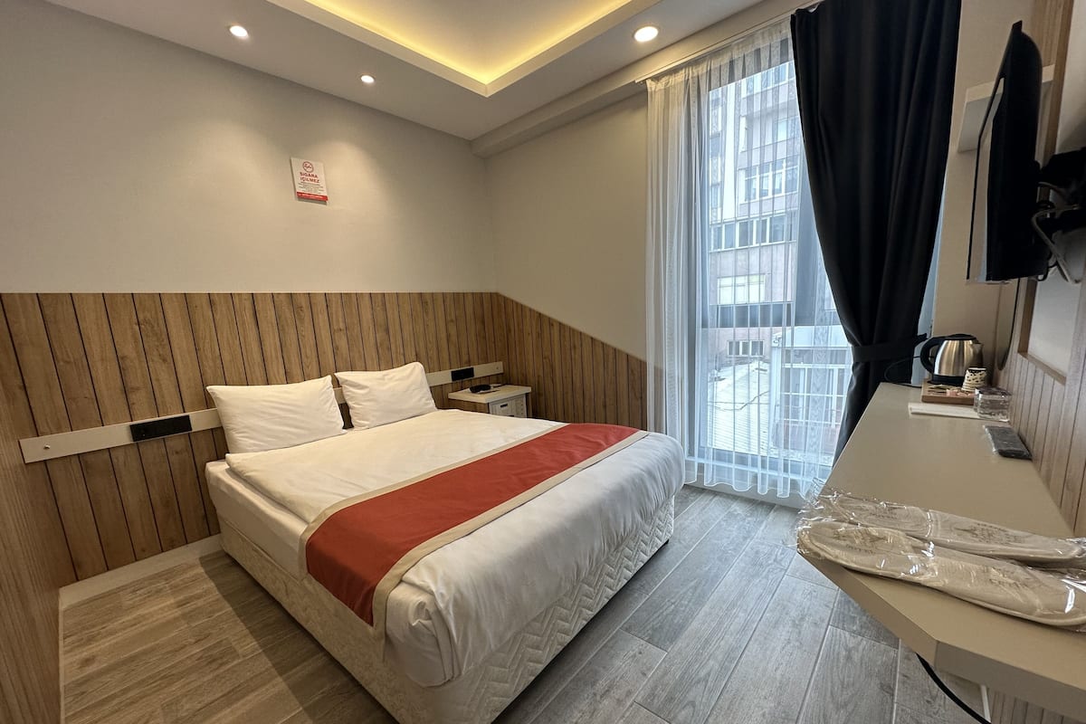 Une chambre d’hôtel avec une tête de lit en bois, un grand lit avec une literie blanche, une table de chevet avec une bouilloire, et une vue sur l’extérieur par une fenêtre ornée de rideaux légers.