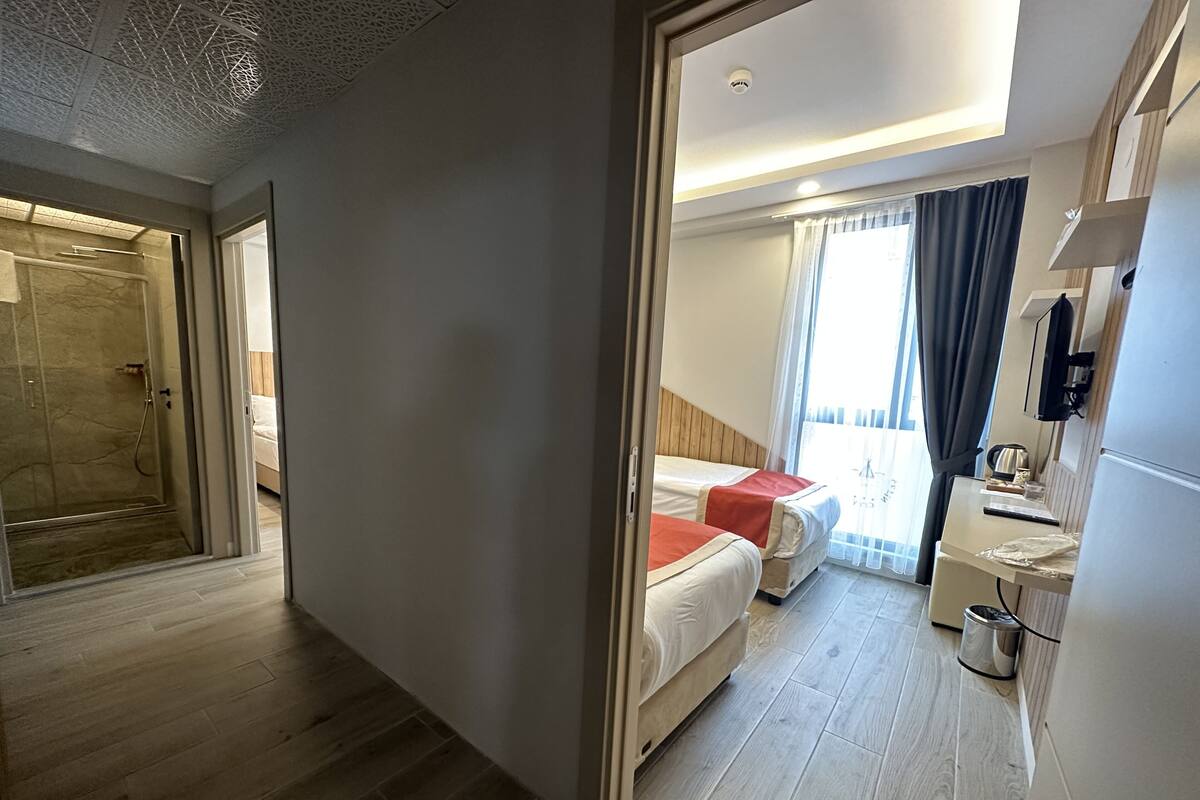 Une chambre d’hôtel avec un plancher en bois, un lit avec une literie rouge et blanche, une table de chevet avec une bouilloire et une fenêtre avec des rideaux.
