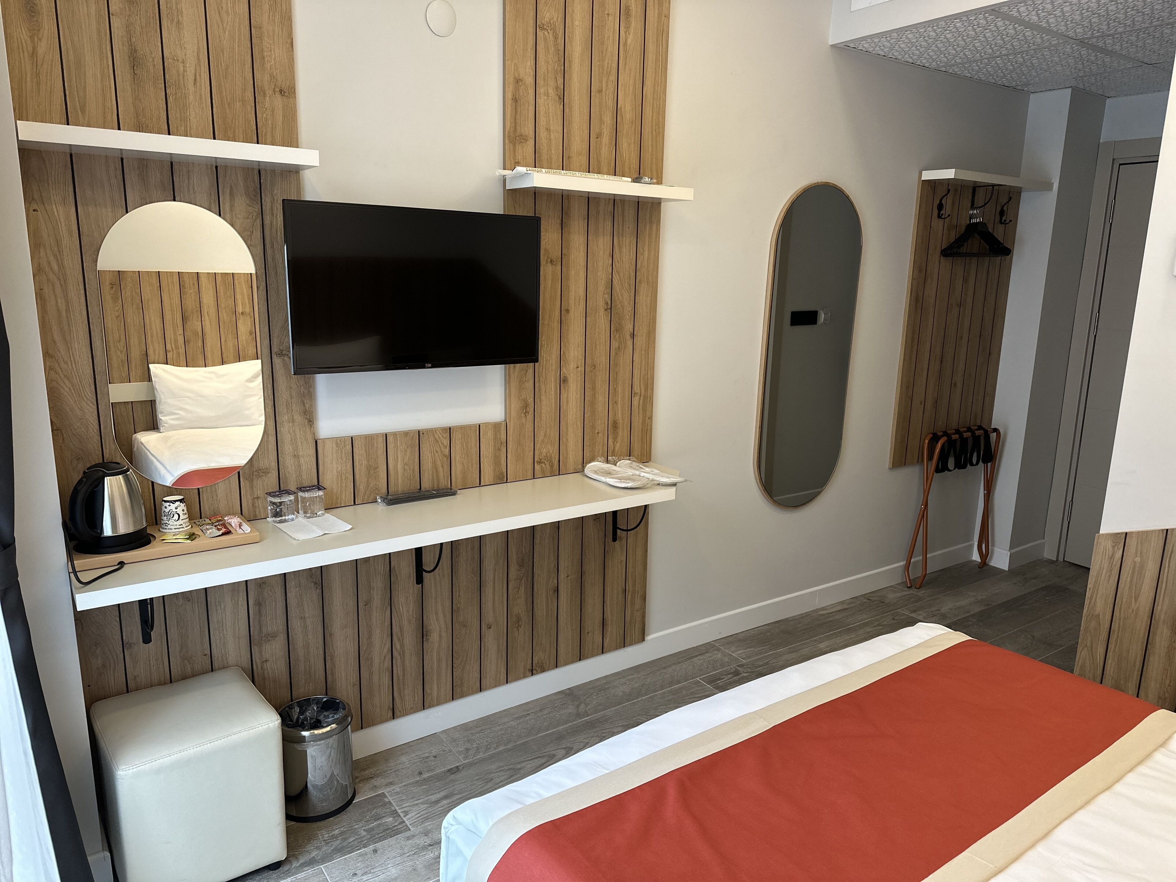 A modern hotel room with a flat-screen TV, a bedside table with a lamp, a mirror, a bed with a red and white bedspread, and a wooden wall paneling.