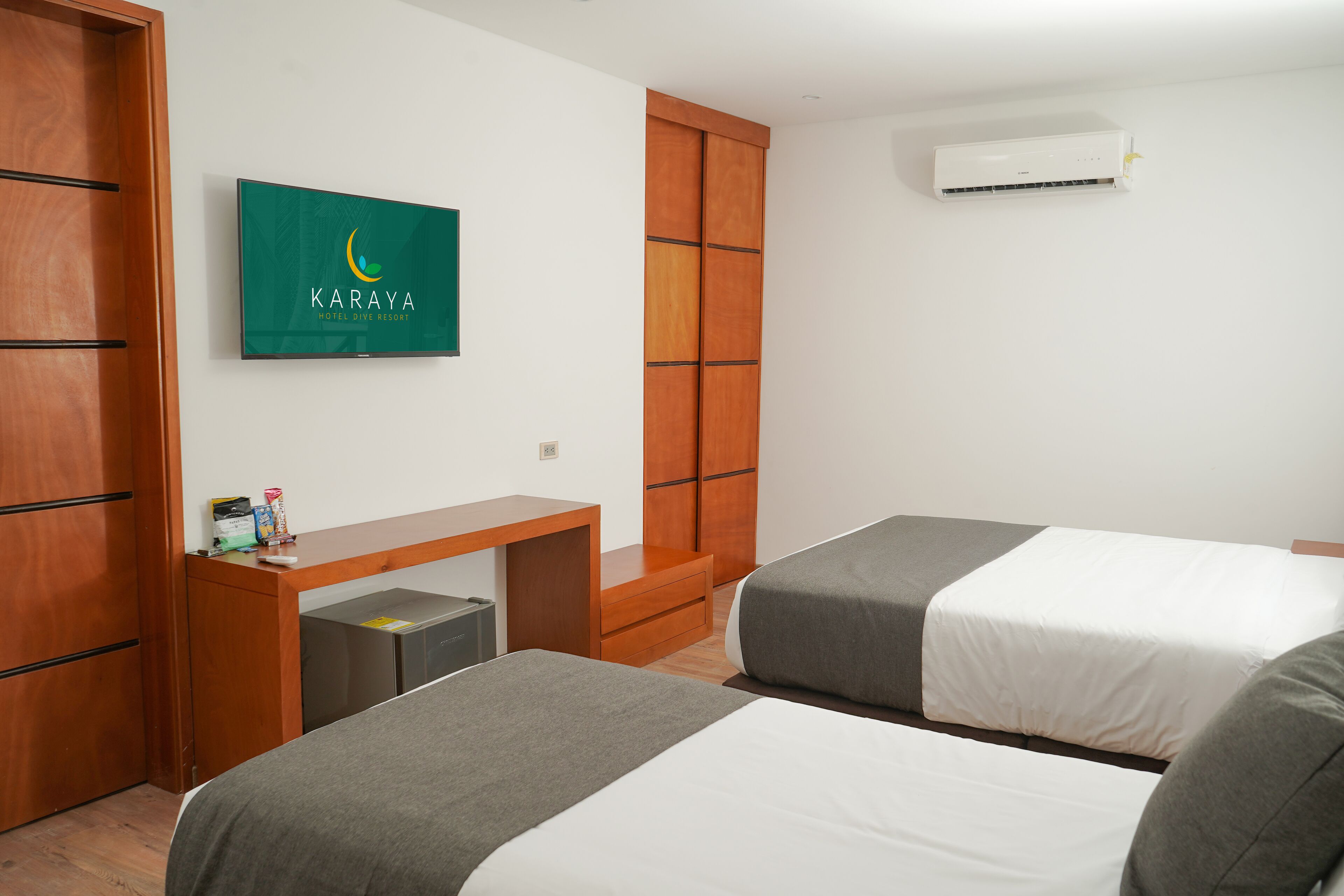 Ein Hotelzimmer mit zwei Betten, einem Holztisch, einer kleinen Kommode, einer Wandklimaanlage und einem gerahmten Schild an der Wand.