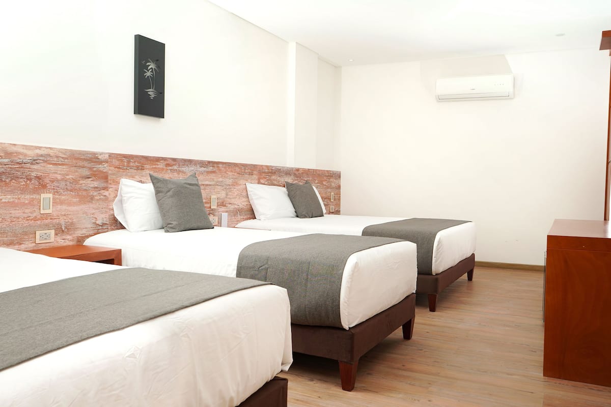 Habitación de hotel con tres camas, cada una con sábanas blancas y mantas grises. Tiene un cabecero de madera, un aire acondicionado instalado en la pared y un televisor de pantalla plana sobre un soporte de madera.