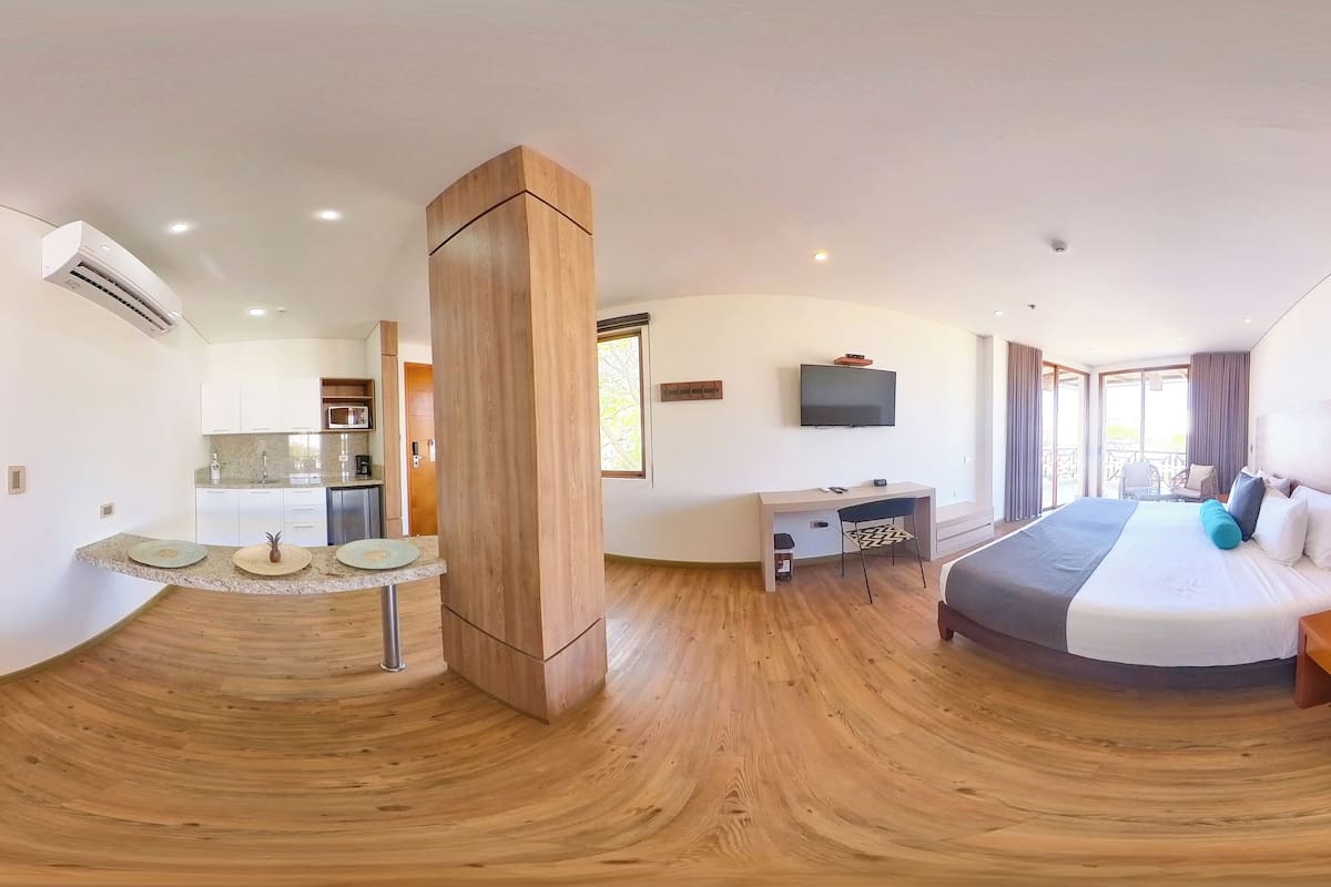 Habitación de hotel moderna con piso de madera curvada, cama con cubrecama gris y blanco, mesita de noche de madera, escritorio con silla, televisor de pantalla plana montado en la pared y un ventanal con cortinas.