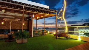 Een terras buiten met kunstgras, houten meubels en een uitzicht op de stad in de schemering.