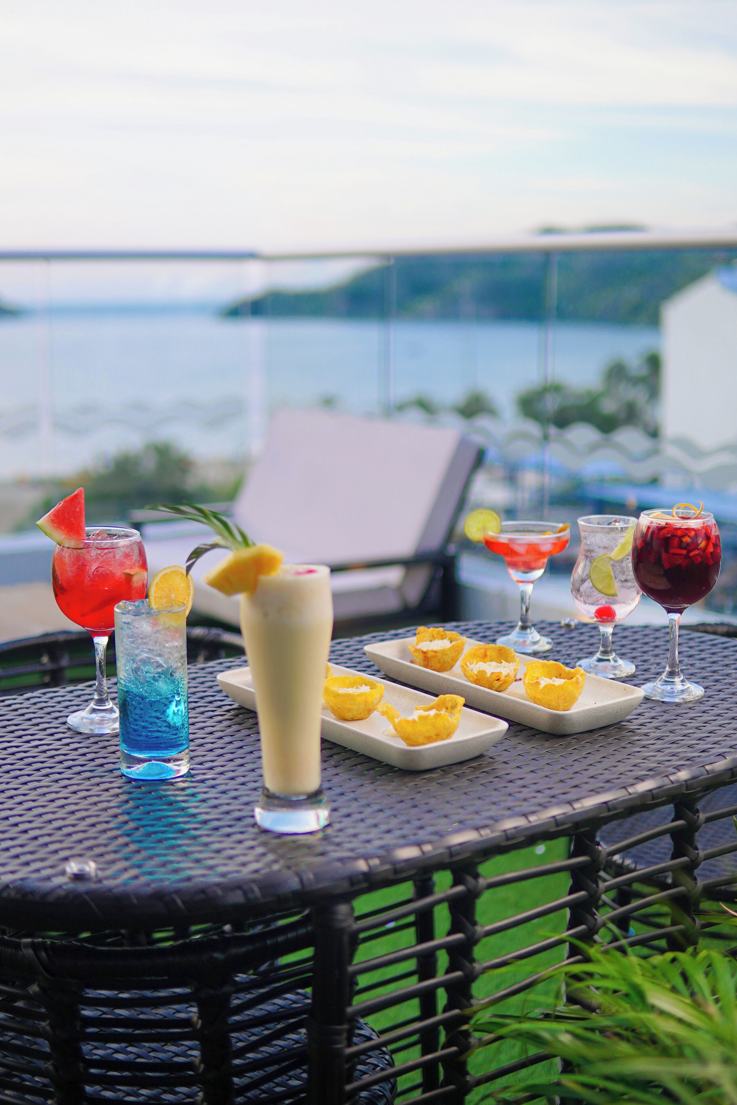 A table with various drinks and appetizers, overlooking a body of water.