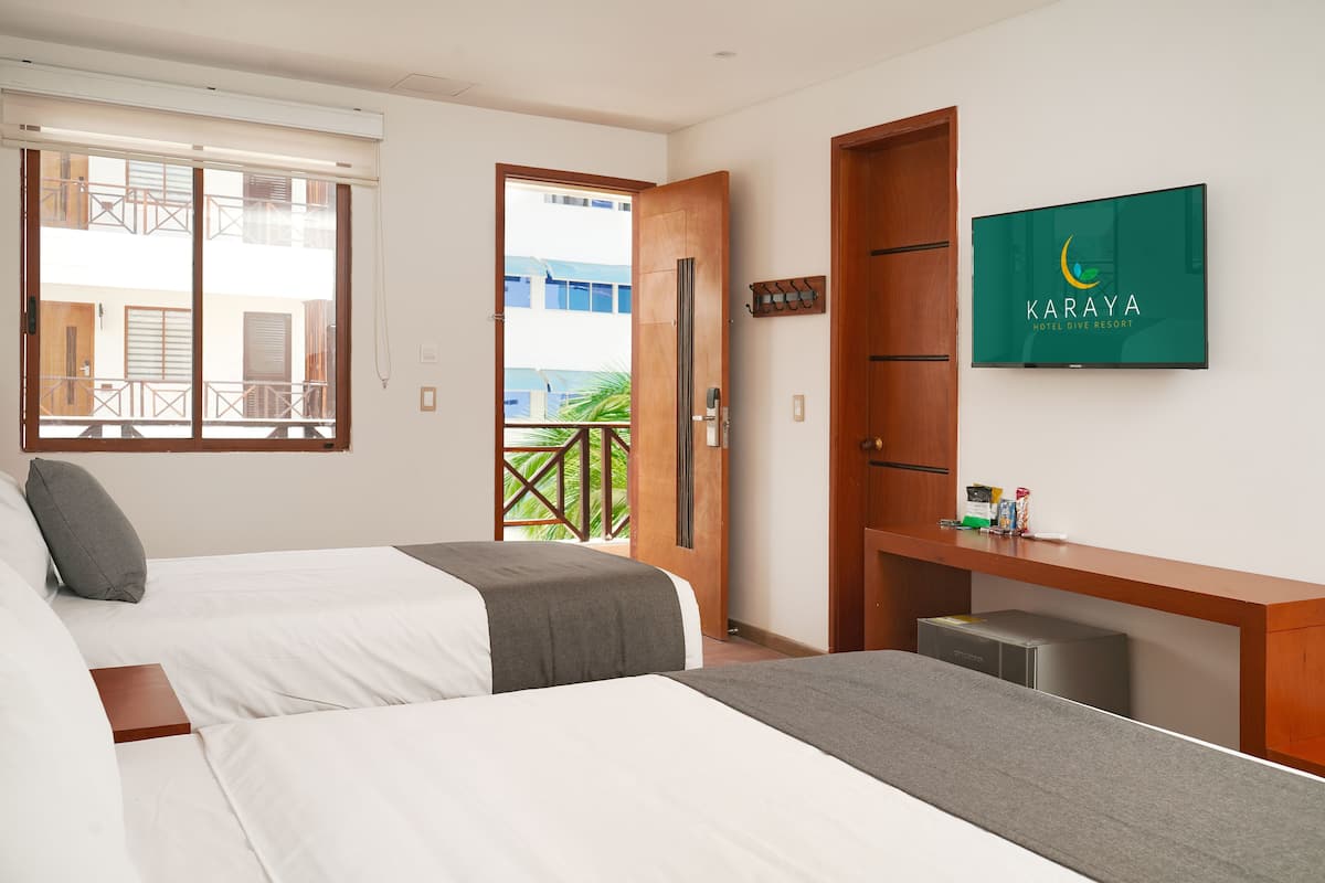 Habitación de hotel con dos camas, un escritorio, un televisor y un balcón con vistas a edificios.