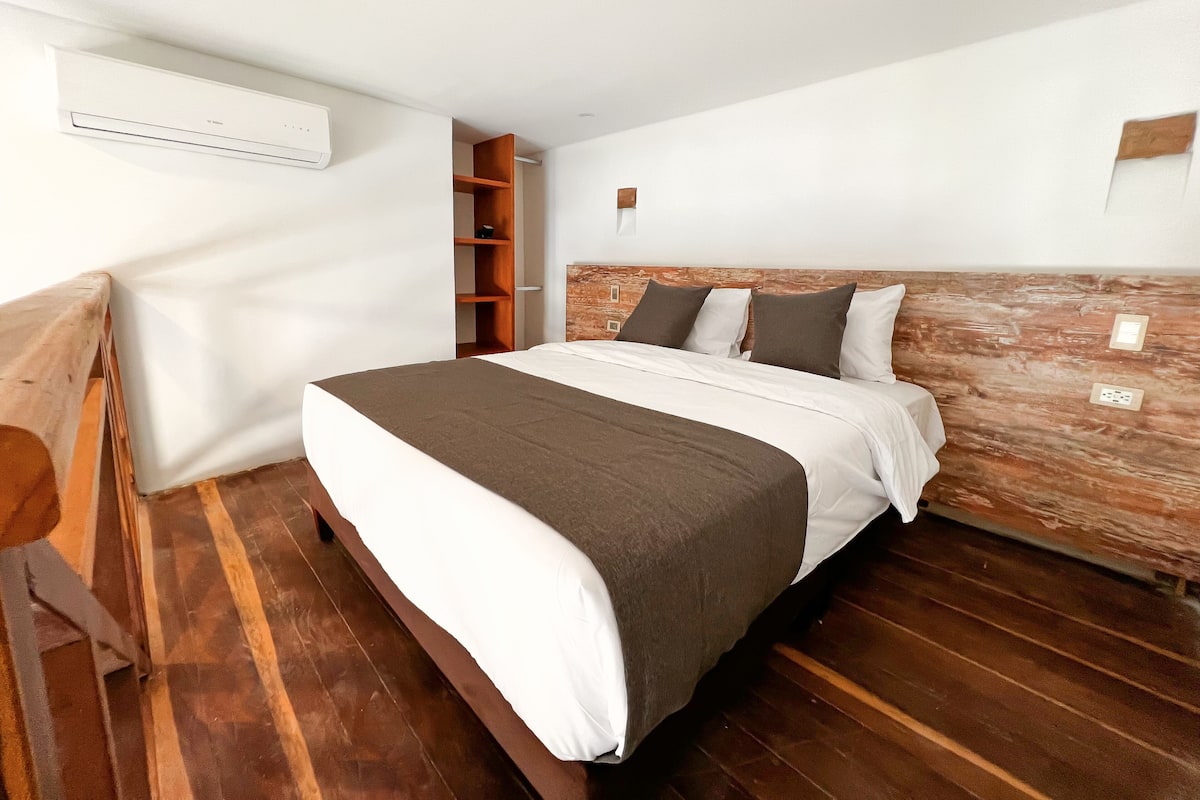 Un dormitorio moderno con cabecera de madera, cama con ropa de cama blanca y marrón, estantería de madera y un equipo de aire acondicionado en la pared.