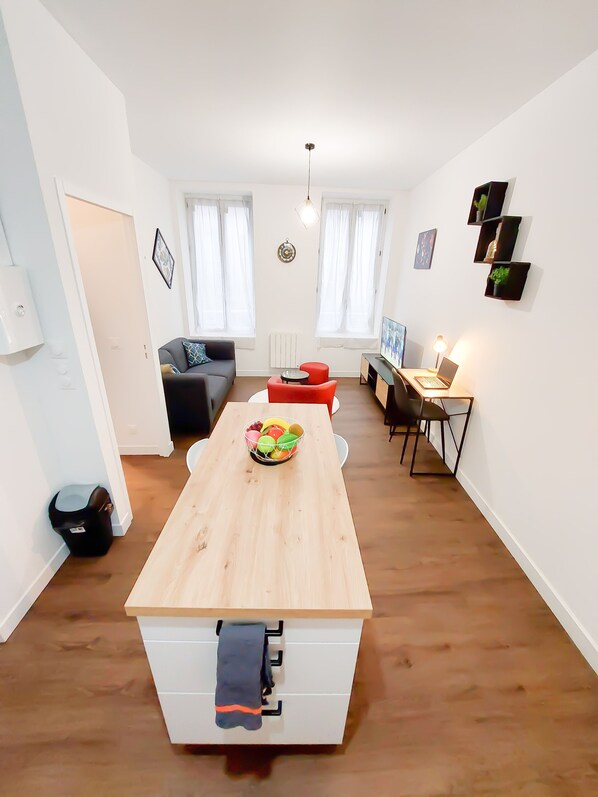Un salon moderne avec une table à manger en bois, un canapé et un fauteuil rouge. On aperçoit un ordinateur portable sur un bureau et une petite table sur laquelle se trouve un saladier rempli de fruits.