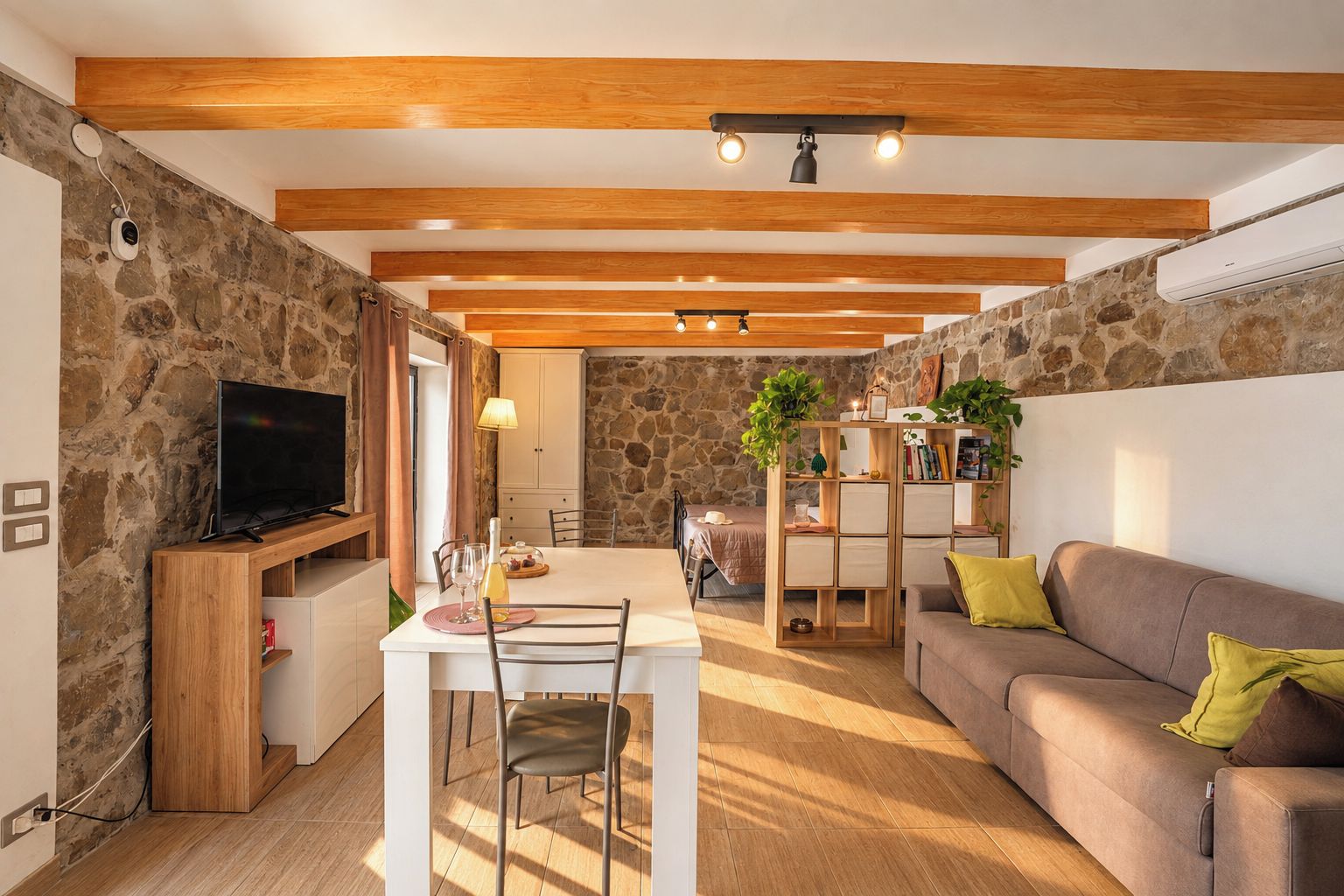 A modern living room with a stone wall, wooden beams, a TV, a dining table with chairs, and a sofa with cushions.