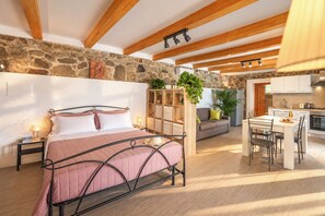 A bedroom with a bed, bedside tables, a stone wall, a wooden ceiling, and a kitchenette in the background.