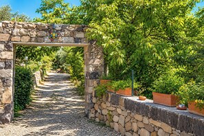 A stone pathway leading through a garden with a stone archway, surrounded by lush greenery and potted plants.