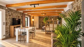 A room with a stone wall, wooden beams, a television, a dining table with chairs, a sofa, and a bookshelf.