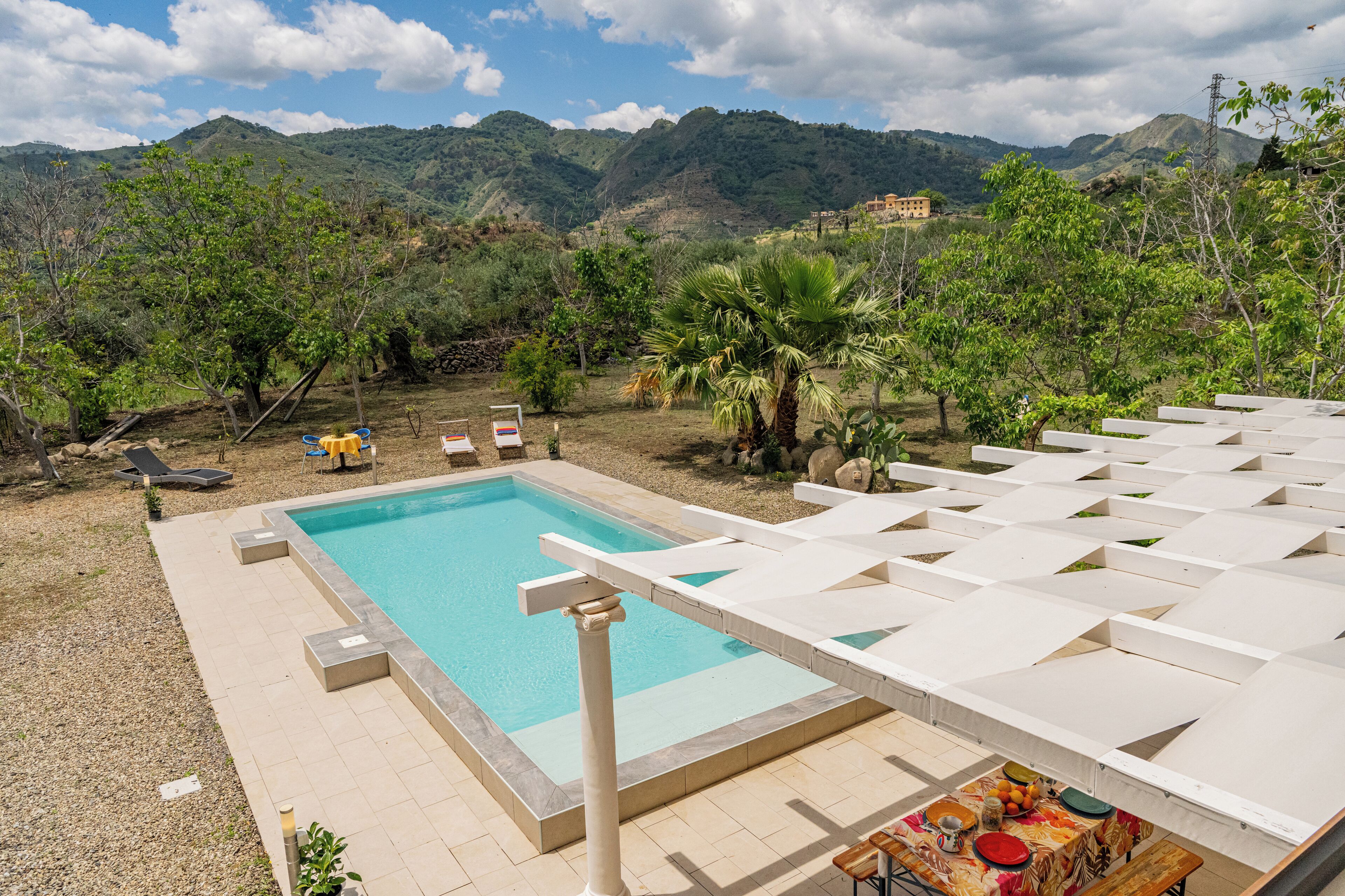 A swimming pool with lounge chairs and a table, surrounded by greenery and mountains in the background.
