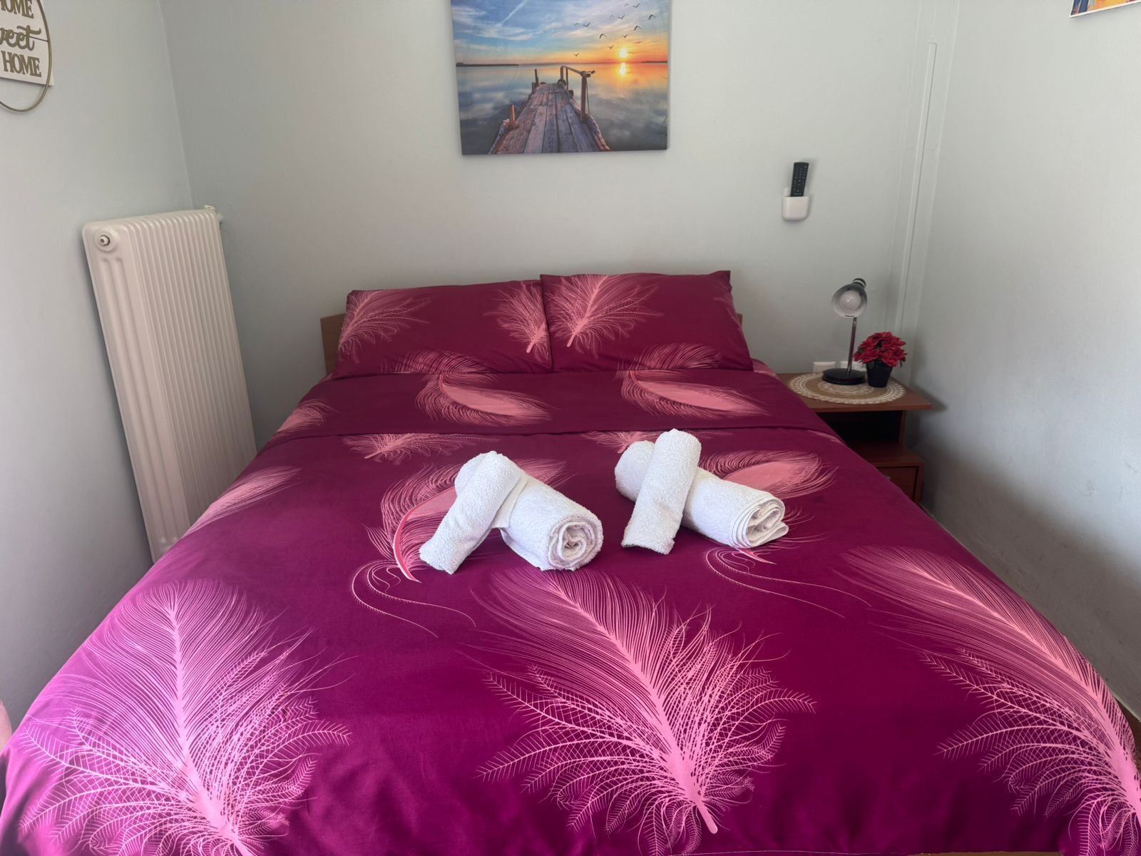 Un letto rifatto con cura, una coperta rosa a motivi pennacchi, due asciugamani arrotolati, un quadro raffigurante un tramonto e un termosifone a sinistra.