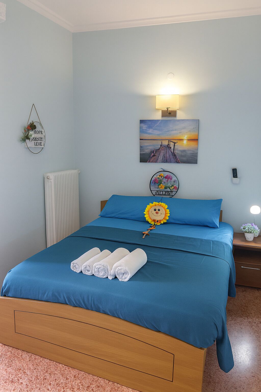 A neatly made bed with blue bedding, a wall-mounted picture of a sunset, a wall clock, and a small plant on the nightstand.