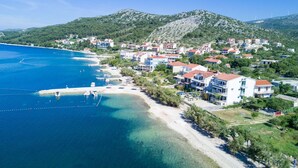 An aerial view of a coastal town with buildings along the beach, clear blue waters, and a hilly backdrop.