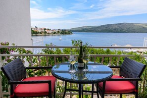 A balcony with a round glass table, two wicker chairs with red cushions, a bottle of white wine, and a glass. The view includes a lake with boats and a distant shoreline. - Luxury apartment literally on the beach - whirlpool and sauna (Seget Vranjica)