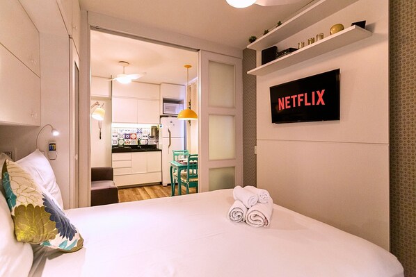 A hotel room with a neatly made bed, a wall-mounted TV displaying "NETFLIX", and a view into the adjacent kitchen area.