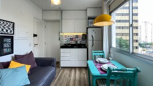 Un espacio habitable compacto con kitchenette, una zona de comedor con mesa y sillas, un sofá con cojines y una ventana con vistas a la ciudad.