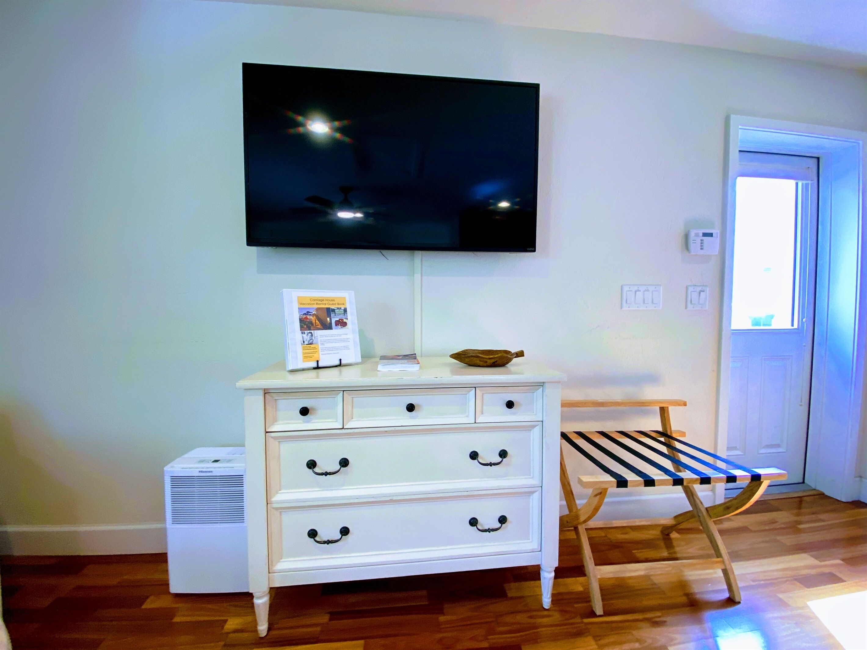 A room with a white dresser, a flat-screen TV mounted above it, a small table with a book, and a wooden bench.