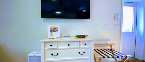 A room with a white dresser, a flat-screen TV mounted above it, a small table with a book, and a wooden bench.
