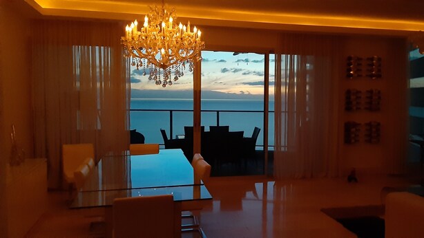 A modern living room with a glass table and chairs, a chandelier, and a view of the sea through large windows.