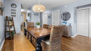 A dining area with a wooden table, wicker chairs, a hanging light fixture, and a wall clock. - 3rd Floor Ocean View, Pools/Hot Tub- Shore Dr (Myrtle Beach)