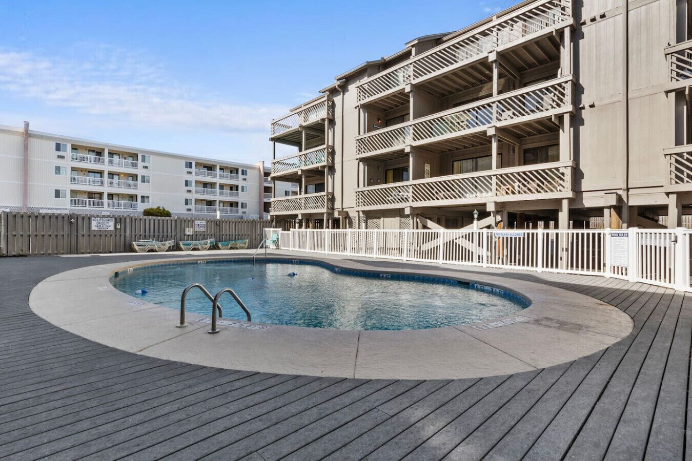 A pool area with a wooden deck, a pool with a ladder, and a building with balconies and a fence.