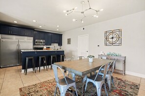 A modern kitchen with dark cabinets, a stainless steel refrigerator, a central island with stools, and a dining table with chairs.