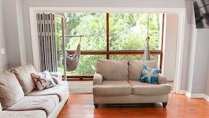 Un salon avec un canapé beige, un coussin décoratif à motifs et un fauteuil suspendu près d’une grande fenêtre donnant sur un paysage verdoyant.