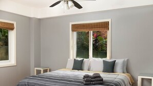 Un dormitorio con cama, dos mesitas de noche, ventilador de techo y ventana con vistas a follaje.