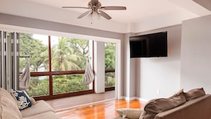 A spacious living room with a large window, a ceiling fan, and a flat-screen TV mounted on the wall.