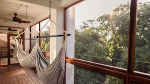 A hammock suspended from ceiling chains in a room with wooden flooring and large windows overlooking a forest.