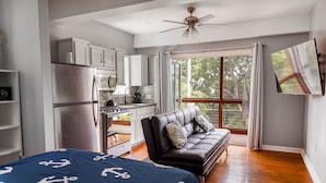A compact bedroom with a bed, a sofa, a kitchenette, and a large window.