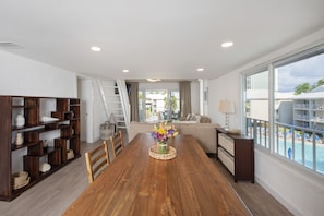 A spacious living area with a wooden dining table, a sofa, and a large window overlooking a pool.