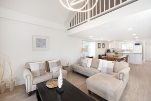 A spacious living area with a modern kitchen in the background, featuring a high ceiling and a staircase.