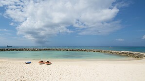 Ein Strand mit klarem türkisblauem Wasser, einem steinernen Wellenbrecher und einigen Booten im Sand.