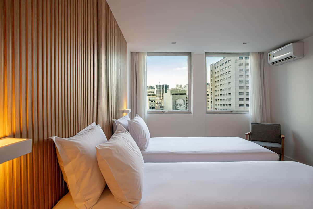 A hotel room with two beds, a wooden panel wall, a window with a city view, and an air conditioning unit.