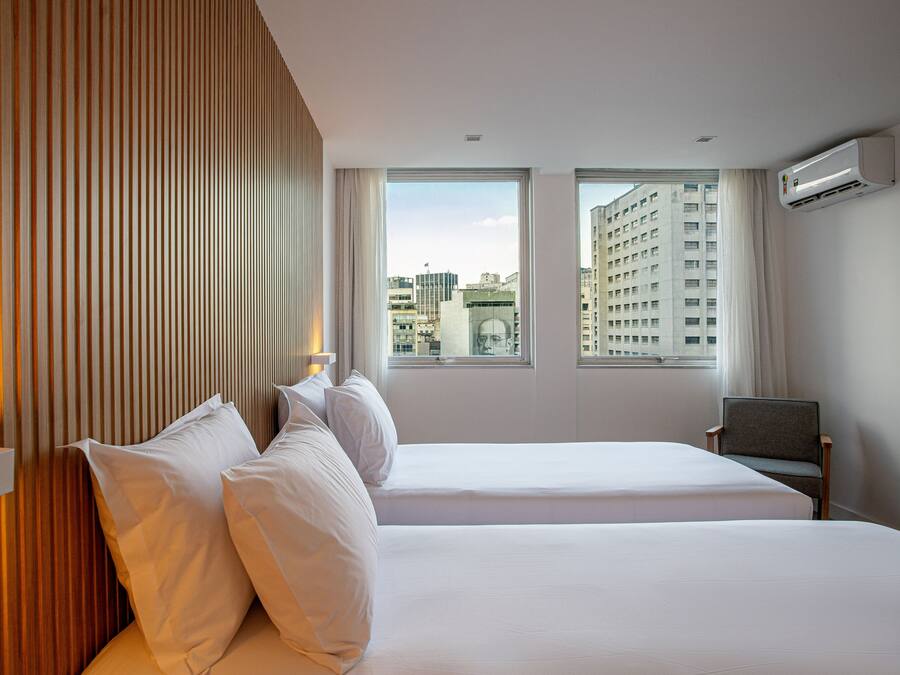 Une chambre d’hôtel avec deux lits, un mur recouvert de panneaux en bois, une fenêtre donnant sur la ville et un appareil de climatisation.