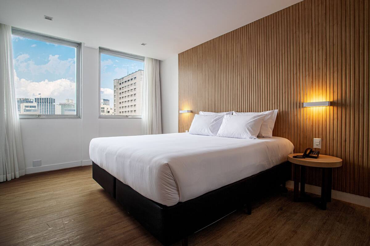 A modern hotel room with a large bed, wooden panel wall, and a view of the cityscape through large windows.
