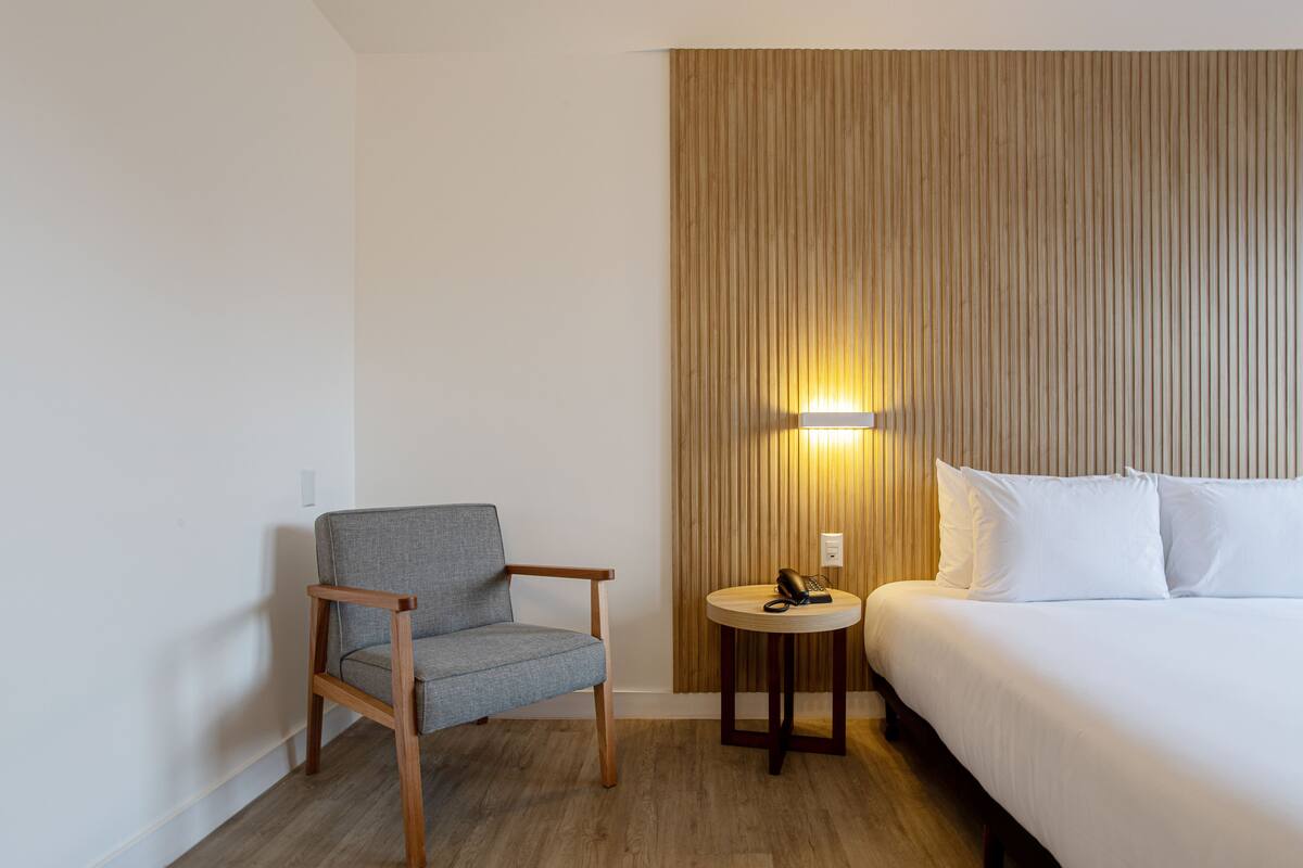A hotel room with a wooden floor, a grey upholstered chair, a round wooden table with a telephone, and a bed with white linens.