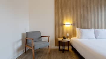 Una habitación de hotel con piso de madera, una silla tapizada de gris, una mesa redonda de madera con un teléfono y una cama con ropa blanca.