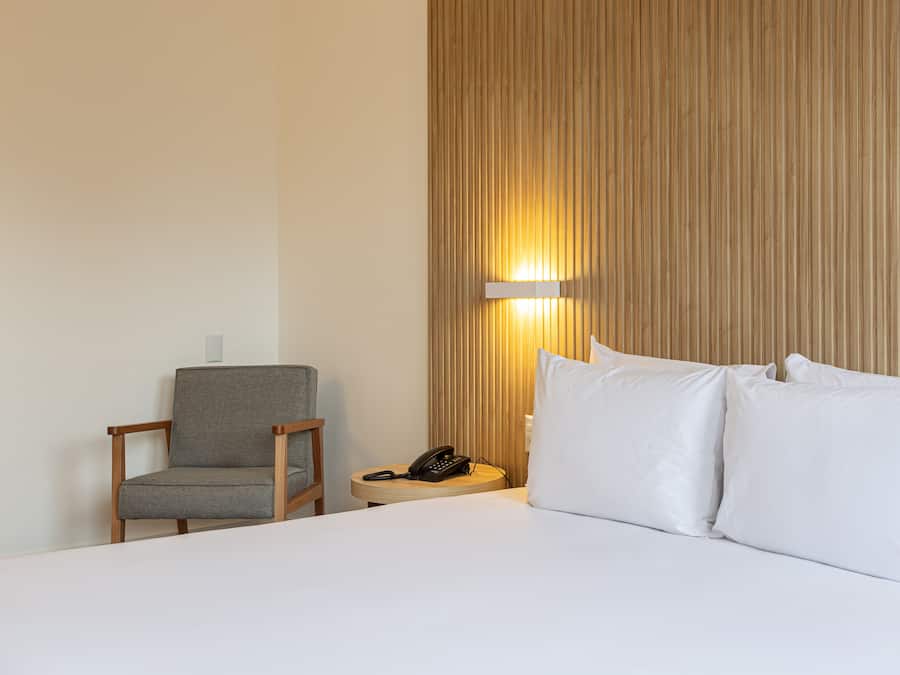 Et hotelværelse med seng, stol, et natbord med telefon og en lampe på væggen.